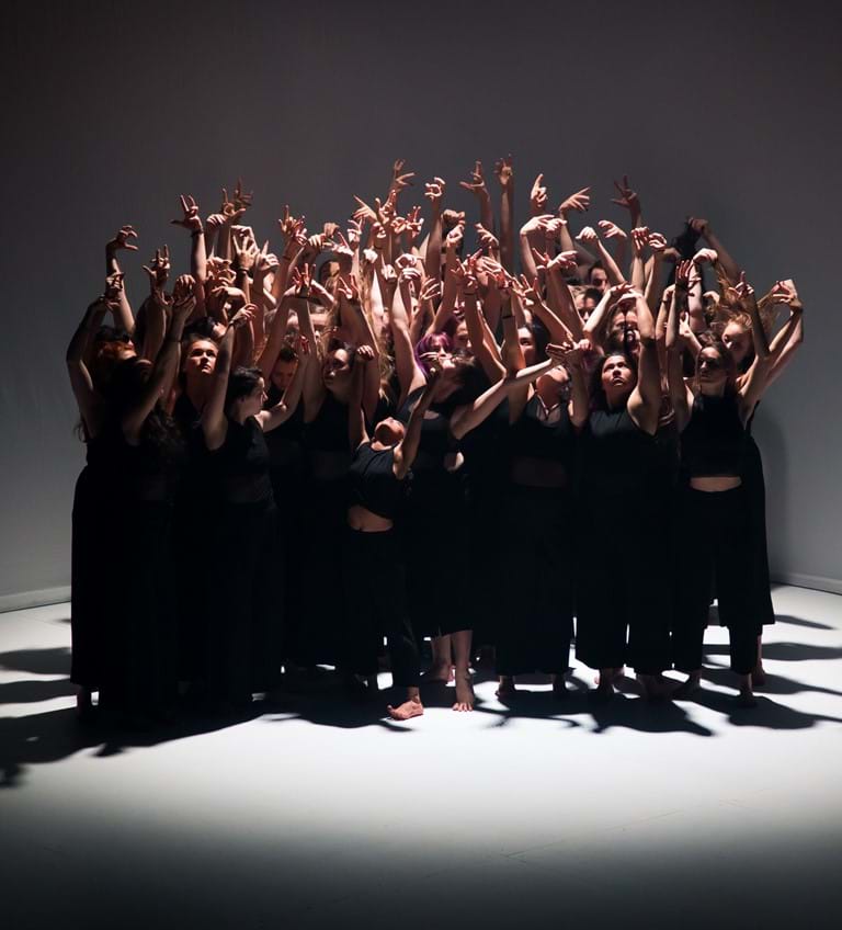 A group of dancers wearing black stand in a spotlight with their arms in the air 